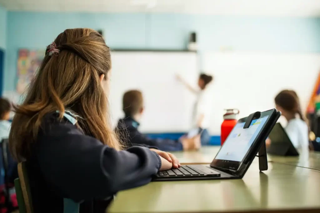 Schoolgirl looking at the teacher who is explaining on the white board while working on her laptop